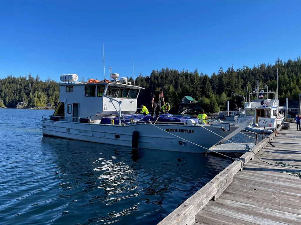 Crew loading and securing supplies on a work vessel docked at a marina, with forested shoreline and calm coastal waters in the background on a clear day.